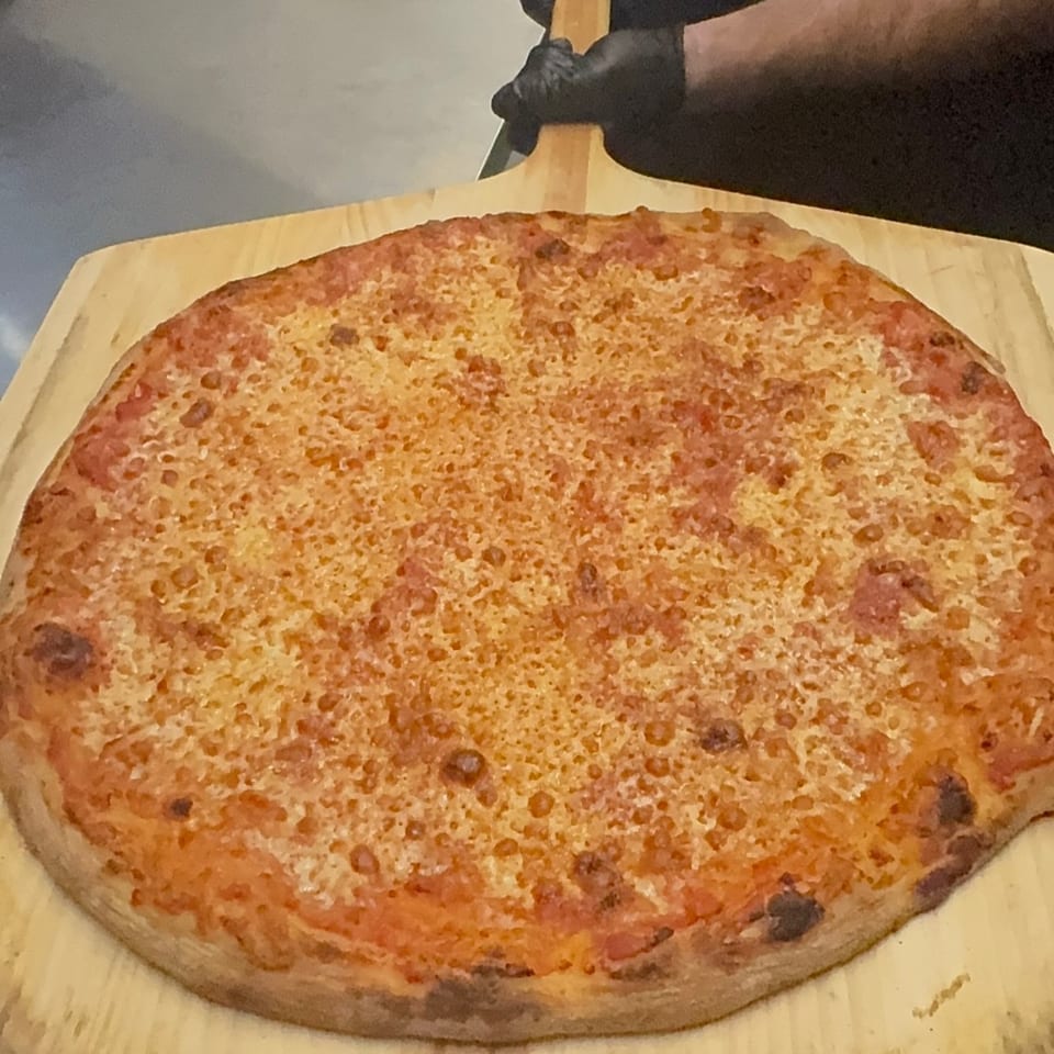 A plain cheese pizza on a wooden peel, held by a person in black gloves.